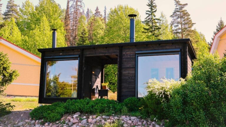 Asiakkaalle toimitettu ulkoasultaan tumma saunamökki kuvattuna ulkoa päin. Mökissä on sauna ja tupa isoilla ikkunoilla ja niiden välistä löytyy mökin halkaiseva katettu terassi.