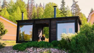 Asiakkaalle toimitettu ulkoasultaan tumma saunamökki kuvattuna ulkoa päin. Mökissä on sauna ja tupa isoilla ikkunoilla ja niiden välistä löytyy mökin halkaiseva katettu terassi.
